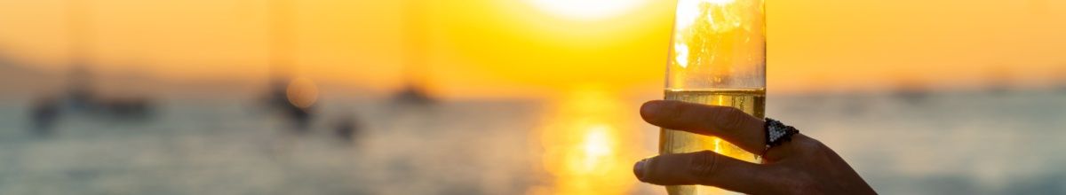 Eine Frauenhand mit einem Ring am Finger hält ein Champagnerglas an einem Strand im Sonnenuntergang.