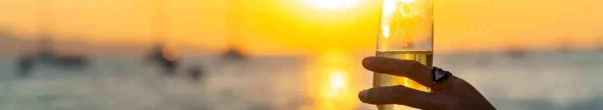 Eine Frauenhand mit einem Ring am Finger hält ein Champagnerglas an einem Strand im Sonnenuntergang.