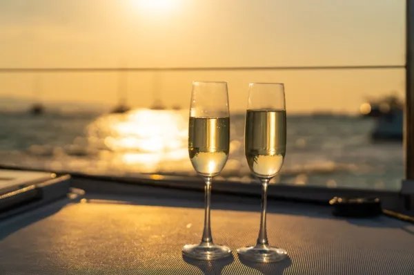 Zwei Sektgläser stehen auf einem Tisch an der Reling des Kreuzfahrtschiffes und im Hintergrund geht die Sonne unter.