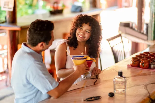 NCL Getränkepakete Philosophie Lifestyle Flotte Ein glückliches Paar stößt an einer gemütlichen Bar mit farbenfrohen Cocktails an.