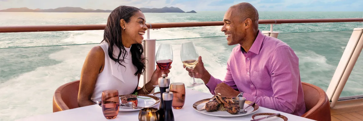NCL Getränkepakete Abendessen Wein Genuss Ein lächelndes Paar stößt beim exklusiven Abendessen auf dem Außendeck eines Schiffes von Norwegian Cruise Line mit Rotwein und Weißwein an.
