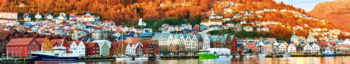 Panoramablick auf den historischen Hafen Bryggen in Bergen, Norwegen, im warmen Licht der tiefstehenden Sonne während des Mittsommers, mit bunten Häusern, Schiffen im Wasser und der Stadt am Berghang.