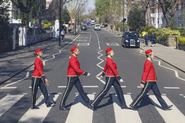 Vier Personen in roten Cunard-Uniformen überqueren im Gänsemarsch den berühmten Zebrastreifen der Abbey Road in London. Die Szene ist eine Nachstellung des ikonischen Beatles-Albumcovers, mit einem Londoner Taxi im Hintergrund.
