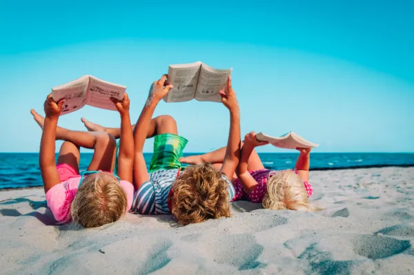 Drei Kinder liegen auf dem Rücken im Sand am Meer und halten jeweils ein offenes Buch über sich, um darin zu lesen. Im Hintergrund sind das blaue Meer und ein klarer Himmel zu sehen.