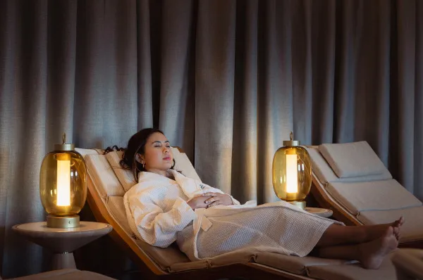 Eine Frau in einem weißen Bademantel entspannt mit geschlossenen Augen auf einer bequemen, gepolsterten Liege in einem Spa-Ruheraum.