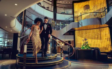 Elegantes Paar auf einer Treppe an Bord eines Cunard-Schiffes Ein elegant gekleidetes Paar, die Frau in einem goldenen Paillettenkleid und der Mann im Smoking, schreitet eine geschwungene, mit Teppich ausgelegte Treppe in einem luxuriösen Innenbereich eines Kreuzfahrtschiffes hinab