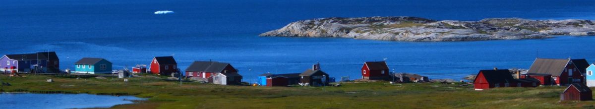 Eine Häuserreihe in Grönlands Tundra umgeben von Wasser und kargen Felsen.