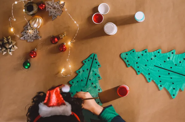 Von oben: Kind mit Weihnachtsmütze bemalt einen Tannenbaum aus Pappe, daneben Farbtöpfe, Kugeln und Lichterkette.