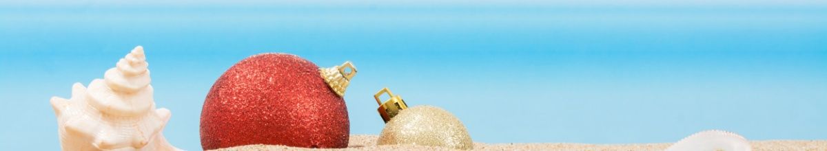 Muscheln und eine rote Weihnachtskugel im hellen Sand vor blauem Himmel.
