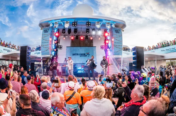 Eine Band spielt live auf einer großen Bühne an Deck eines Kreuzfahrtschiffs, umgeben von einer jubelnden Menge von Passagieren, viele davon verkleidet oder mit Accessoires, die eine ausgelassene Party-Stimmung vermitteln.
