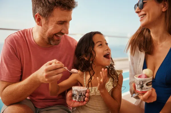 Ein Mann füttert ein Kind, das begeistert lacht, mit einem Löffel Eis. Eine Frau schaut den beiden lächelnd zu, während die Familie gemeinsam an Deck eines Schiffes sitzt und eine sonnige Auszeit genießt.