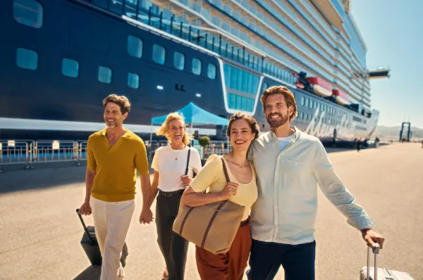 Zwei glücklich aussehende Paare laufen mit ihrem Gepäck an einem sonnigen Hafenkai. Im Hintergrund liegt ein großes, blaues Kreuzfahrtschiff, das auf ihre Einschiffung wartet.
