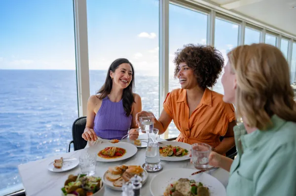 Drei Frauen sitzen lachend an einem Esstisch in einem Restaurant auf einem Kreuzfahrtschiff. Durch die großen Fenster blicken sie auf das offene Meer, während sie sich unterhalten und eine Mahlzeit genießen.