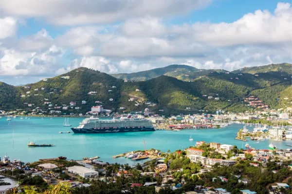 Ein Mein Schiff Kreuzfahrtschiff ankert in einer malerischen Bucht mit türkisblauem Wasser. Im Hintergrund erheben sich grüne, dicht bewachsene Hügel und an der Küste ist eine kleine Hafenstadt zu sehen.
