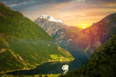 Ein Kreuzfahrtschiff, dass einen Fjord entlangfährt umgeben von bewachsenen hohen Bergen mit einer sinkenden Sonne im Hintergrund.