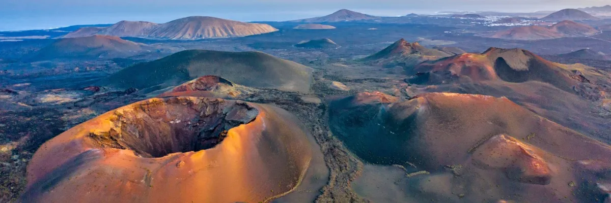 Blick über die Krater der Vulkanlandschaft im Nationalpark Timanfaya auf Teneriffa. Darüber ein von Cirruswolken bedeckter Himmel mit einer hinter den Wolken versteckten Sonne.