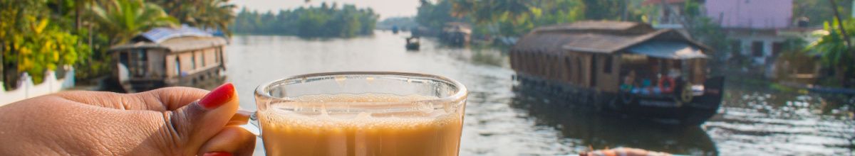 Eine Hand mit roten Fingernägeln hält ein Glas mit einem Getränk (möglicherweise Tee oder Kaffee) vor einer Wasserlandschaft mit traditionellen Hausbooten und üppiger Ufervegetation im Hintergrund. Das Bild fängt eine ruhige Reisestimmung ein.