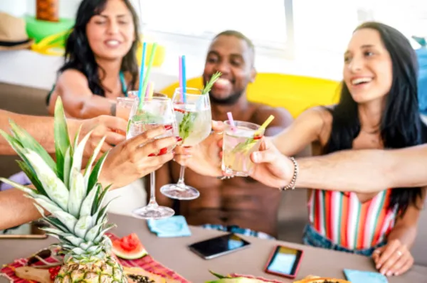 Gruppe von Freunden stößt mit Cocktails an. ine Gruppe fröhlicher, junger Erwachsener stößt gemeinsam mit verschiedenen Cocktails und Getränken an einem Tisch an. Im Vordergrund ist eine Ananas zu sehen, die Szene wirkt sommerlich und gesellig.