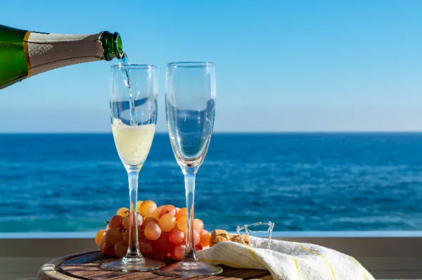Sekt wird in Gläser mit Meerblick eingegossen Sekt oder Champagner wird aus einer Flasche in ein Sektglas gegossen, daneben steht ein weiteres Glas und eine Traube Weintrauben. Im Hintergrund ist das weite, blaue Meer unter klarem Himmel zu sehen, was eine luxuriöse Urlaubsatmosphäre schafft.