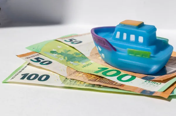 Spielzeugschiff auf Euro-Geldscheinen Ein blaues Spielzeugschiff liegt auf einem Stapel aus 50- und 100-Euro-Scheinen vor einem weißen Hintergrund. Das Bild symbolisiert die Kosten, die mit einer Kreuzfahrt oder Schiffsreise verbunden sind.