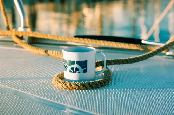Tasse mit Schiffssteuerrad-Logo auf Bootsdeck mit Tau Eine weiße Tasse mit dem blauen Logo eines Steuerrads steht auf dem strukturierten Deck eines Bootes, umgeben von einem aufgewickelten Tau. Das warme Licht und die maritimen Details erzeugen eine ruhige, nautische Stimmung.