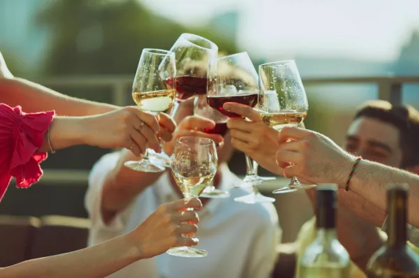 Hände stoßen mit Rot- und Weißweingläsern an Eine Gruppe von Personen stößt mit Weingläsern an, die mit Rot- und Weißwein gefüllt sind. Die Szene findet in einer hellen, geselligen Umgebung statt, möglicherweise im Freien.