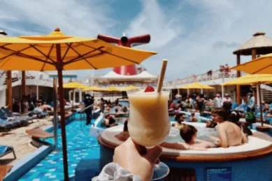 Nahaufnahme eines Cocktails, der vor dem Hintergrund eines belebten Pooldecks auf einem Kreuzfahrtschiff gehalten wird. Das Foto vermittelt die entspannte Atmosphäre und den Genuss, der mit einer Kreuzfahrt und möglicherweise einem Getränkepaket verbunden