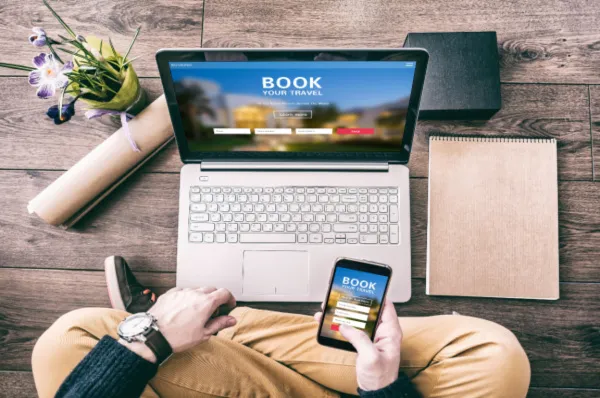 Kreuzfahrt online buchen am Laptop und Smartphone Draufsicht einer Person, die auf einem Holzboden sitzt und über einen Laptop sowie ein Smartphone mit geöffneten Buchungsmasken eine Reise online bucht.