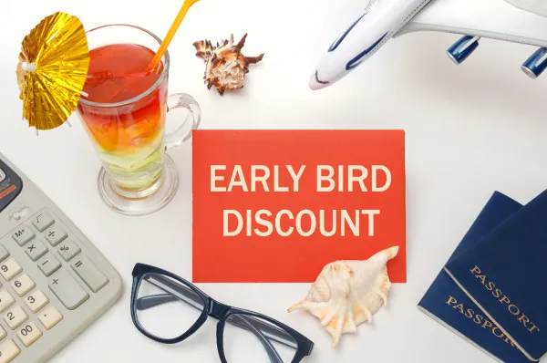 Frühbucher Rabatt für Kreuzfahrt sichern Schreibtisch mit Reiseutensilien wie Pass, Flugzeugmodell, Cocktail und einem Schild mit der Aufschrift "Early Bird Discount", symbolisch für Frühbuchervorteile bei Reisen.