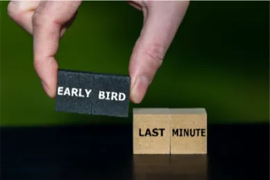 Zwei Finger die einen Holzklotz mit der Aufschrift Early Bird neben einen Klotz legen auf dem Last-Minute steht.