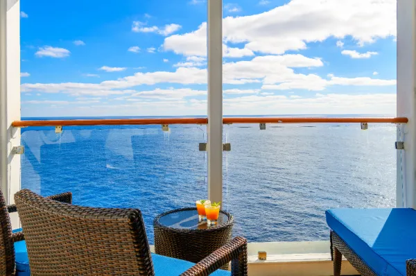 Möblierter Balkon einer Kreuzfahrtkabine mit zwei Stühlen, Tisch und Getränken, der einen weiten Blick auf das blaue Meer bietet. Steht für Kabinen-Komfort, den man eventuell durch Stornokabinen günstiger bekommt.