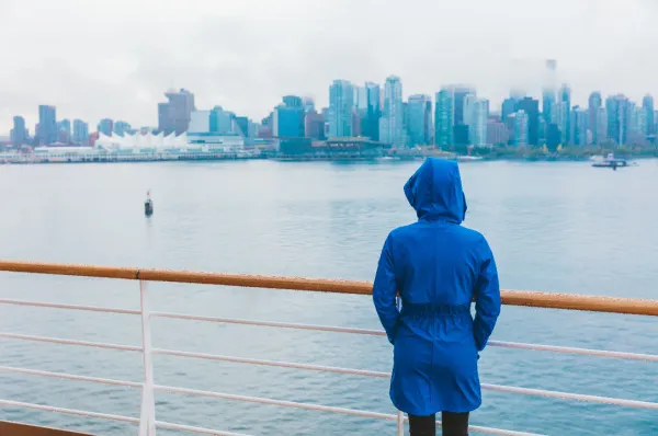Ein Frau in einer blauen Regenjacke an einer Reling, während Sie über die Hafenkante einer Großstadt mit Hochhäusern schaut.
