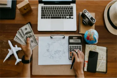 Draufsicht auf einen Schreibtisch für die Reiseplanung mit Laptop, Weltkarte, Kamera und Händen, die Geld und einen Taschenrechner halten, als Symbol für die Budgetierung einer Kreuzfahrt.