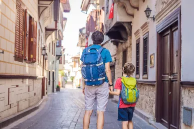 Vater und Sohn mit Rucksäcken erkunden händchenhaltend eine malerische Gasse während eines Landausflugs.
