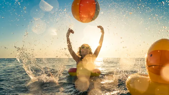 Ein glückliches Kind in einem Quietscheentchen-Schwimmring spielt im Meer mit einem Ball.