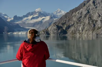 Eine Frau in roter Winterjacke stehend an einer Reling mit Bergen und einem Fjord im Hintergrund.