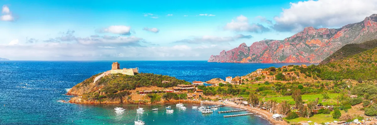 Panoramaaufnahme einer mediterranen Bucht auf Korsika mit einer historischen Festung auf einem grünen Hügel, weißen Segelyachten im türkisfarbenen Wasser und schroffen, roten Felsformationen im Hintergrund.
