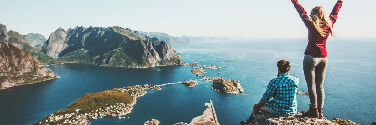 Zwei Personen auf einem steilen Felsgipfel in Norwegen mit einem weiten Panoramablick über die Lofoten-Inseln, Brückenverbindungen und das tiefblaue Nordmeer unter klarem Himmel.