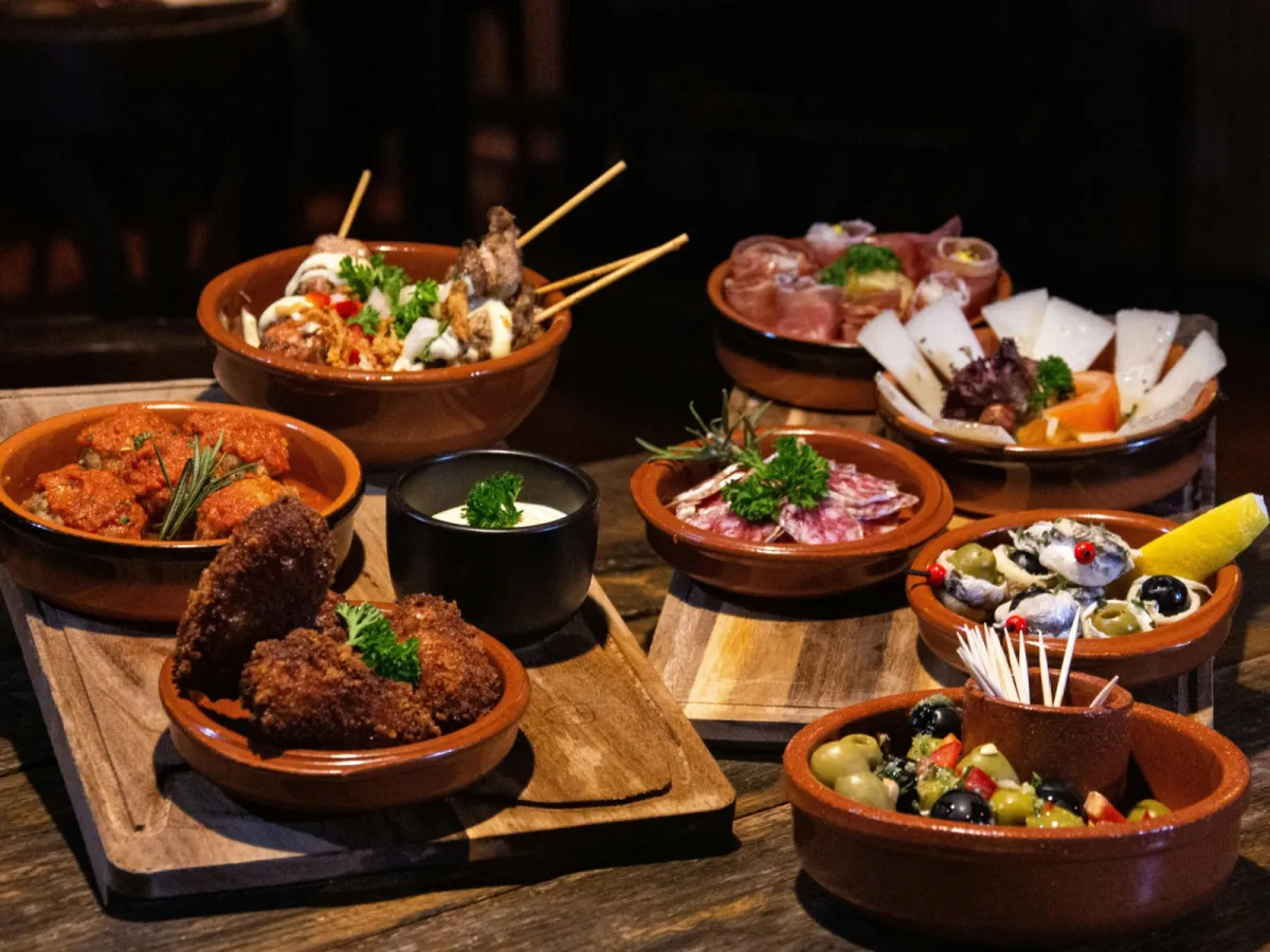 Traditionelle mallorquinische Tapas-Variationen Eine reichhaltige Auswahl spanischer Tapas, serviert in traditionellen Terrakotta-Schalen auf rustikalen Holzbrettern. Zu sehen sind Fleischspieße, Albondigas in Sauce, Kroketten, Serrano-Schinken, Manchego-Käse, Salami und Oliven.