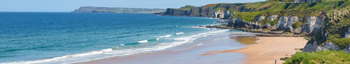 Ein Sandstrand in der Nähe von Portrush, mit blauem Meer und grünen Steilküsten bei Tag.
