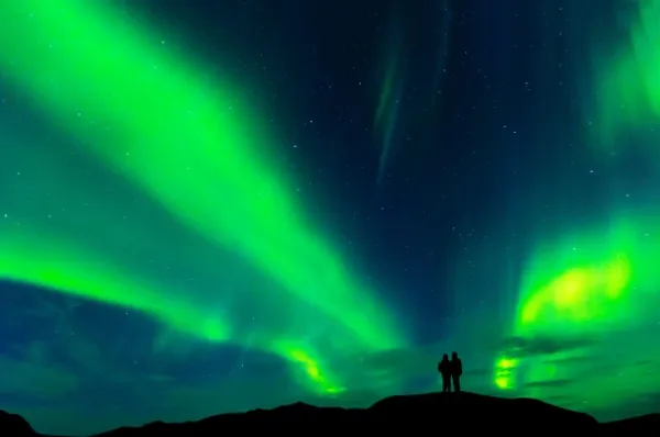 Zwei Personen stehen auf einem Hügel bei Kirkenes und schauen auf die spektakulären, grünen Polarlichter am Himmel.