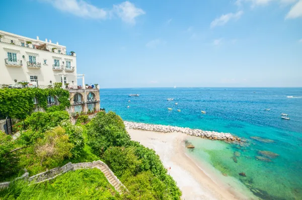 Ein Strand und das klare, azurblaue Meer bei Capri mit einer Villa links im Bild bei Tag.
