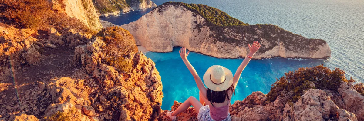 Eine Frau sitzt mit jubelnd erhobenen Armen auf einer hohen Klippe über einer türkisfarbenen Bucht in Griechenland – ein Highlight der mediterranen Routen innerhalb der AIDA Angebote für Alleinreisende.