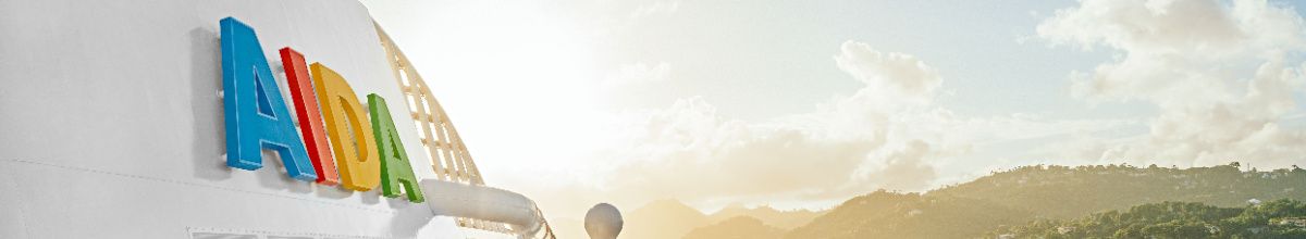 Das ikonische, bunte AIDA Logo an der strahlend weißen Bordwand eines Schiffes vor einer tropischen Küstenlandschaft im sanften Sonnenlicht – passend zu den exklusiven AIDA Angeboten für Alleinreisende.