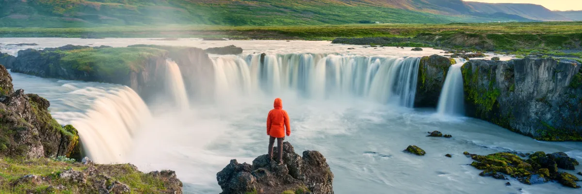 Ein Solo-Reisender in leuchtend roter Outdoor-Jacke betrachtet einen imposanten, breiten Wasserfall in Nordeuropa – ein Symbol für die Freiheit und Naturerlebnisse der AIDA Angebote für Alleinreisende.