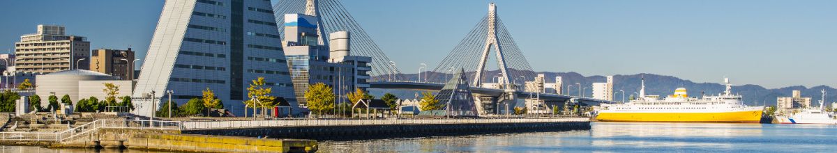 Der Hafen von Aomori, gesäumt mit Hochhäusern und einer Brücke im Hintergrund, sowie einem Kreuzfahrtschiff bei Tag.