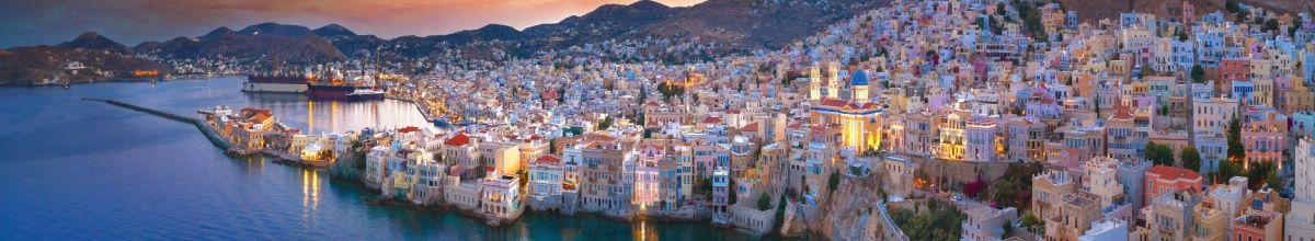 Die Insel Syros und ihre pastellfarbenen Häuser bei Abendlicht am Meer.