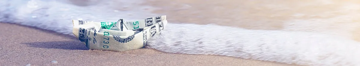 Ein gefaltetes Papierschiff aus einer Banknote an einem Strand.
