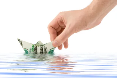 Artwork eines Schiffes gebastelt aus einer 100-Dollar-Banknote mit einer Hand die es auf Wasser setzt.