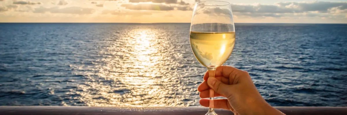 Finden Sie den perfekten Tarif für Ihre Winterkreuzfahrt mit AIDA Eine Hand hält ein Glas Weißwein an der Reling eines Kreuzfahrtschiffes. Im Hintergrund ist der Sonnenuntergang über dem Meer zu sehen.